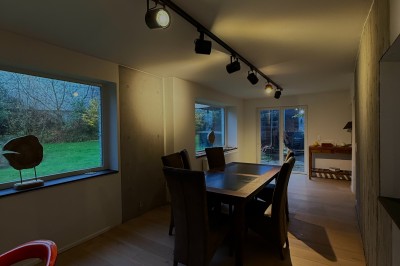 Salle lumineuse avec parquet clair, table et chaises au centre, escalier blanc, machine à coudre ancienne et tableau au mur.