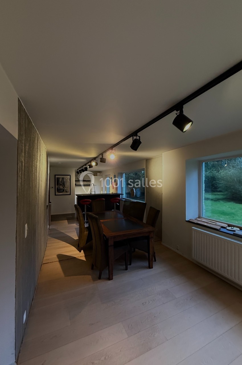 Salle à manger avec table en bois, chaises, éclairage sur rail au plafond et grandes fenêtres donnant sur un jardin.