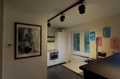 Salle lumineuse avec parquet clair, table et chaises au centre, escalier blanc, machine à coudre ancienne et tableau au mur.