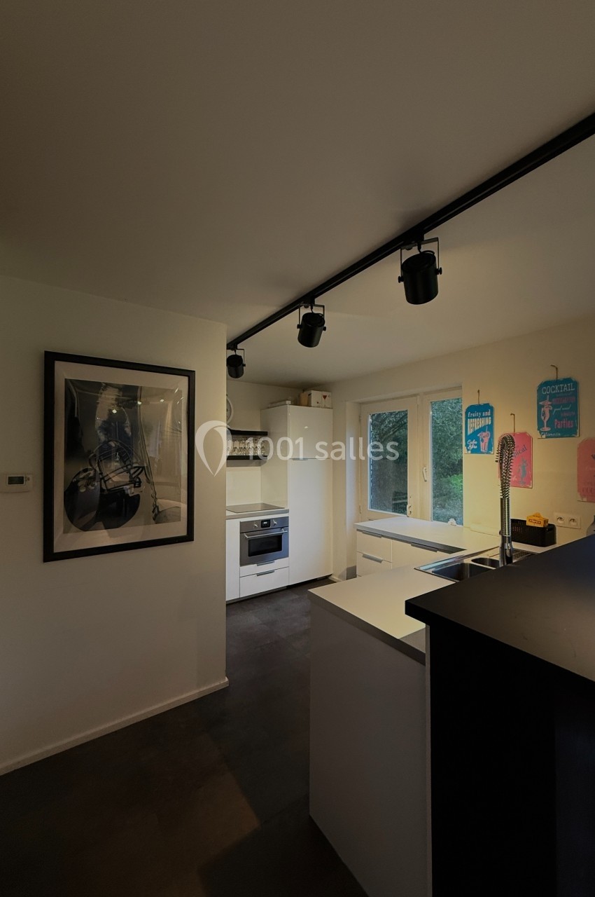 Cuisine moderne avec éclairage sur rail, plan de travail central, électroménagers blancs et fenêtre donnant sur un jardin.