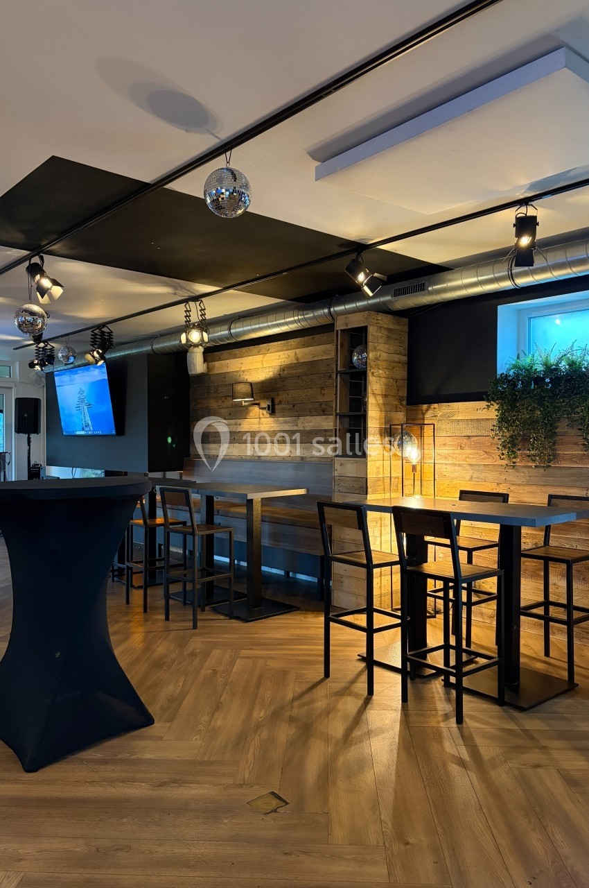 Salle moderne avec parquet, tables hautes, murs en bois, plantes suspendues et éclairage tamisé.
