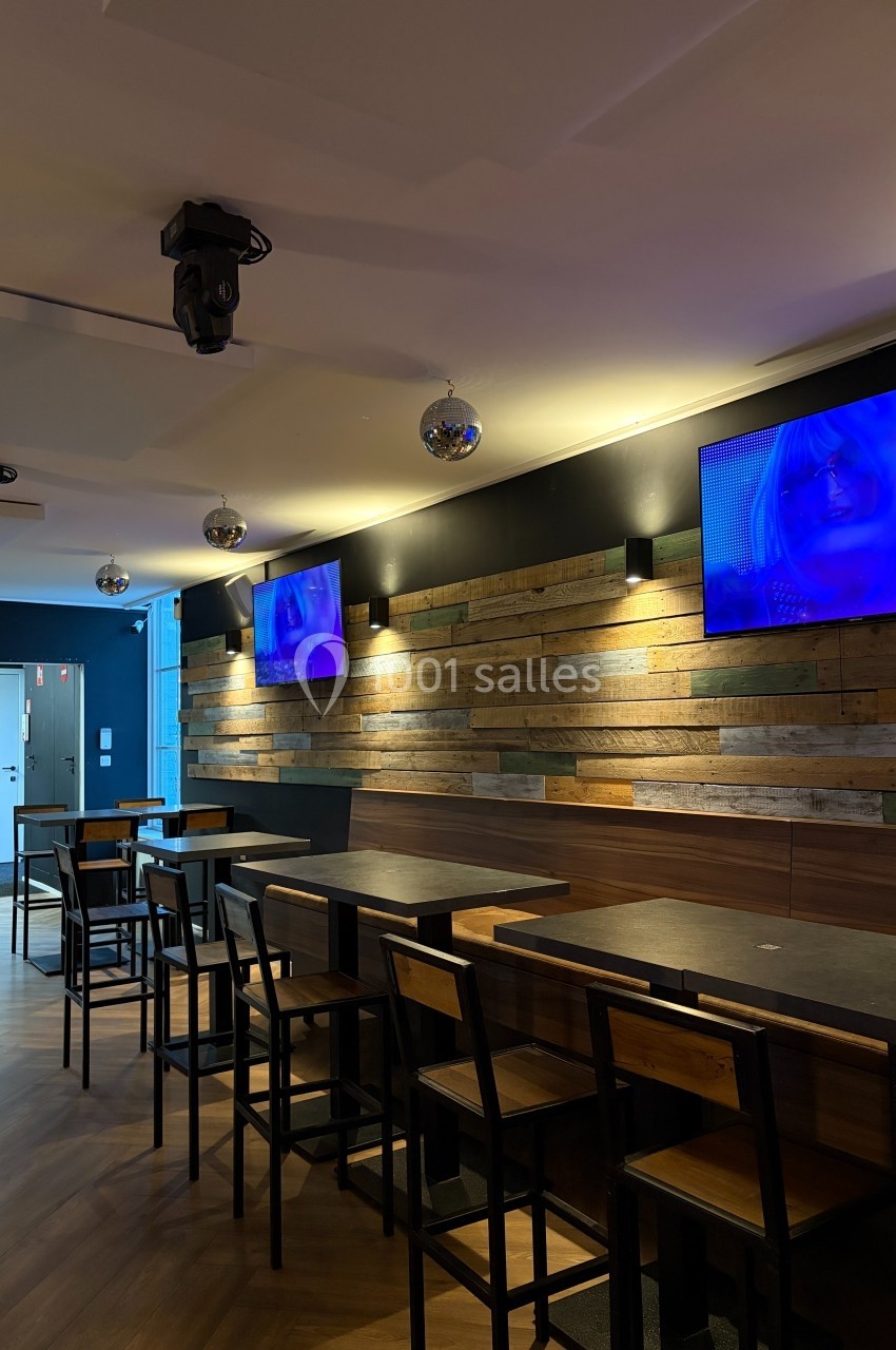 Salle de bar moderne avec tables en bois, chaises noires, écrans allumés et murs décorés de planches en bois.