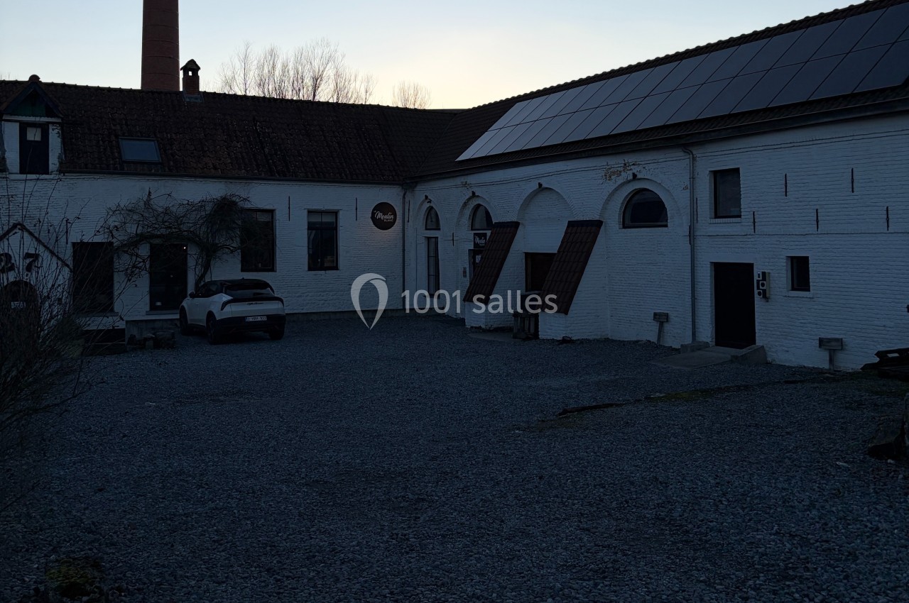 Cour d’un bâtiment blanc avec toit en tuiles et panneaux solaires, une voiture stationnée sur un sol en gravier.