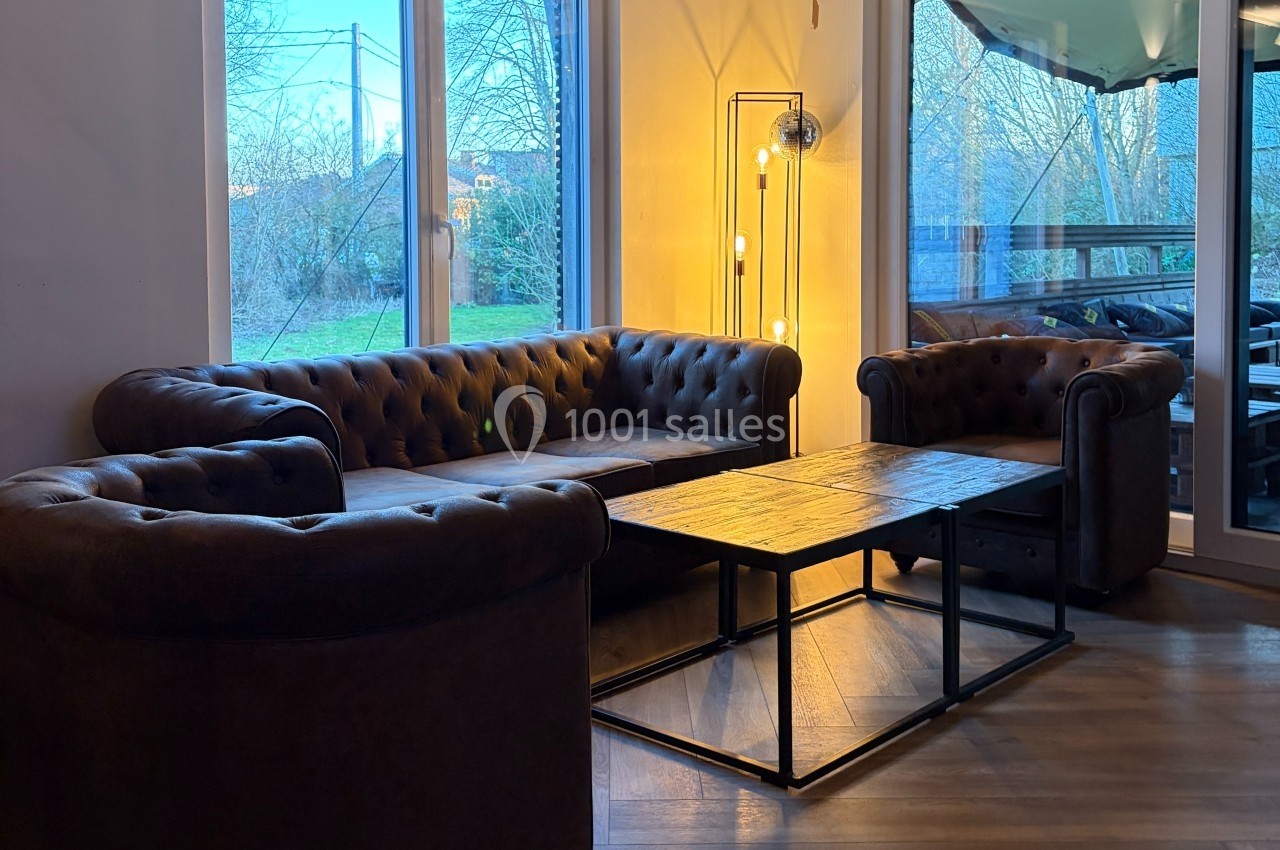 Salon avec deux fauteuils et un canapé en cuir marron autour d'une table basse en bois, éclairé par une lampe et la lumière…