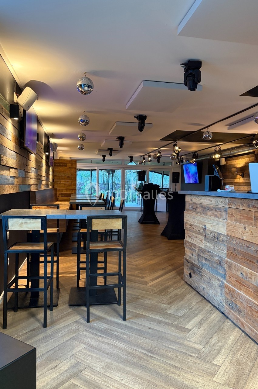Salle moderne avec parquet, murs en bois, tables hautes, bar en bois et éclairage tamisé, vue sur l'extérieur.