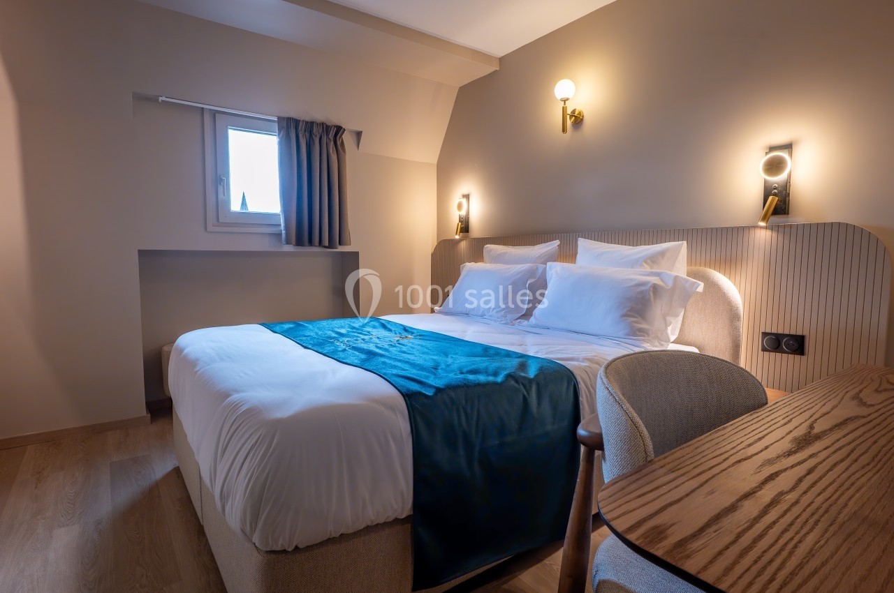 Chambre lumineuse avec lit double, drap bleu, bureau en bois et éclairage mural, fenêtre avec rideau.