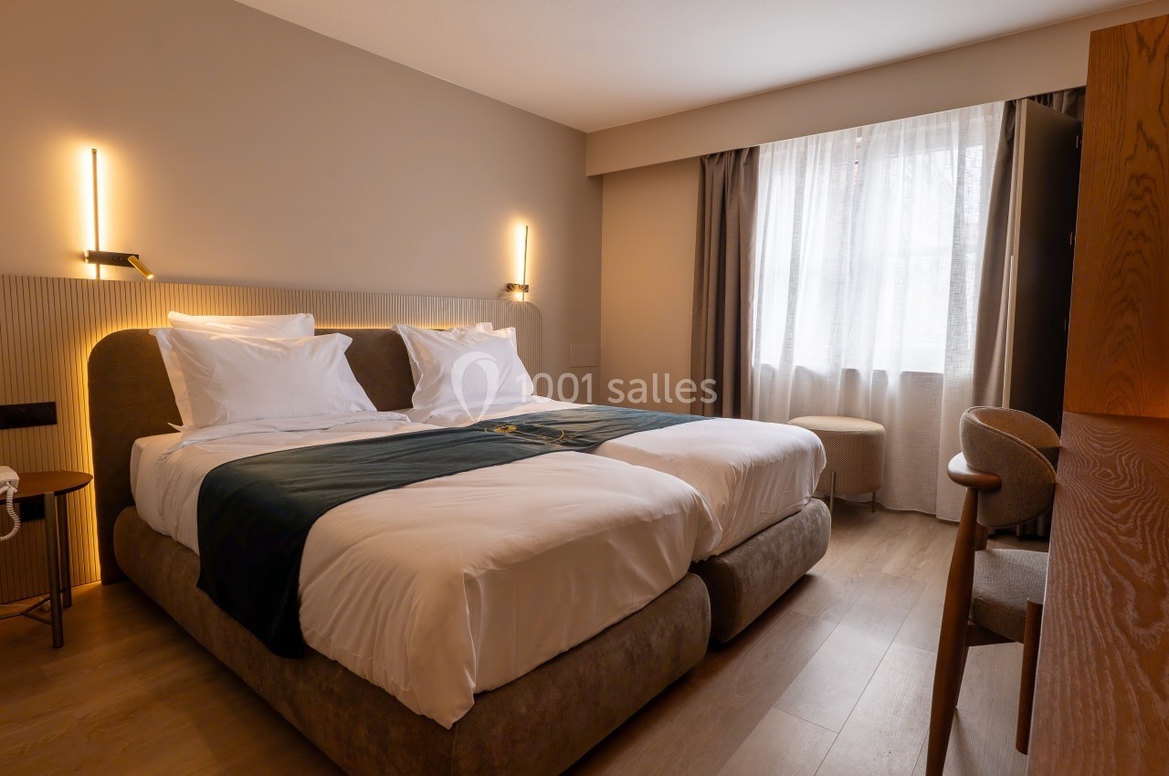 Chambre d'hôtel avec deux lits simples rapprochés, éclairage mural, grande fenêtre avec rideaux et mobilier en bois.