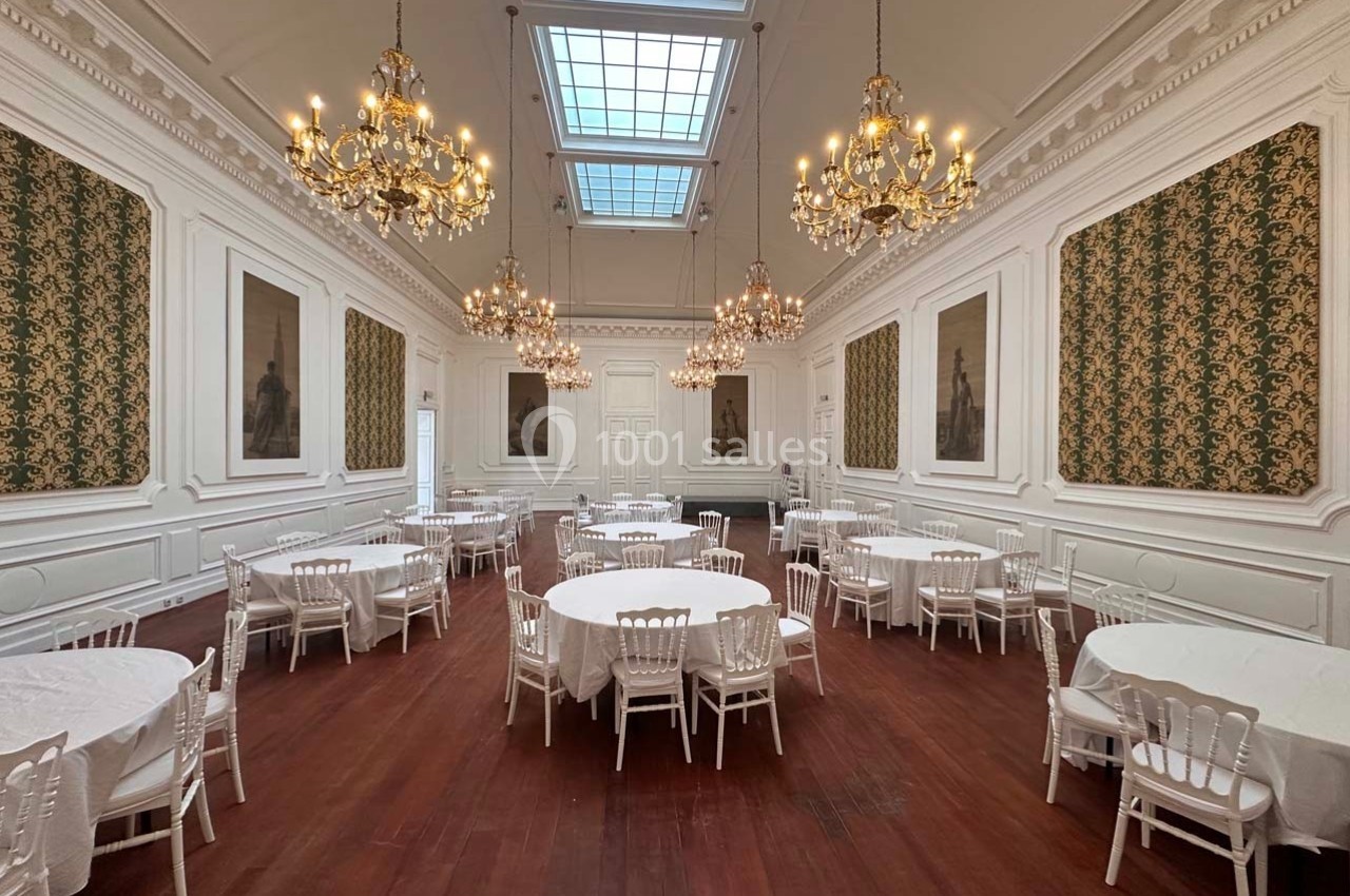Salle élégante avec parquet, tables rondes dressées, chaises blanches, lustres dorés et murs ornés de tableaux et tentures.