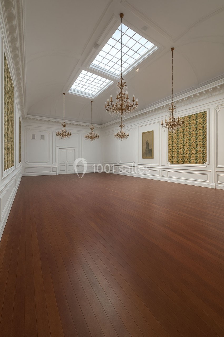 Salle lumineuse avec parquet en bois, murs blancs ornés de moulures, lustres suspendus et tableaux accrochés.