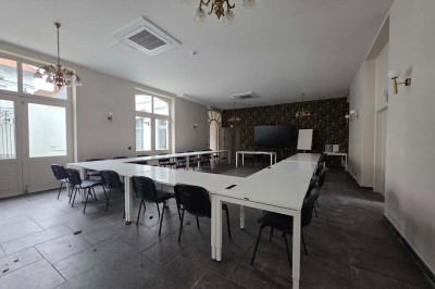 Salle de réunion lumineuse avec de longues tables blanches, chaises noires, écran mural et tableau blanc.
