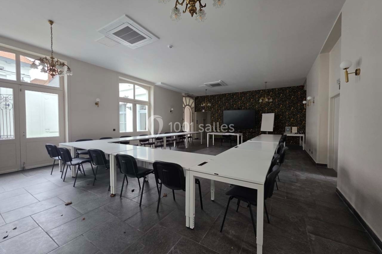Salle de réunion lumineuse avec tables blanches disposées en U, chaises noires et écran de présentation au mur.