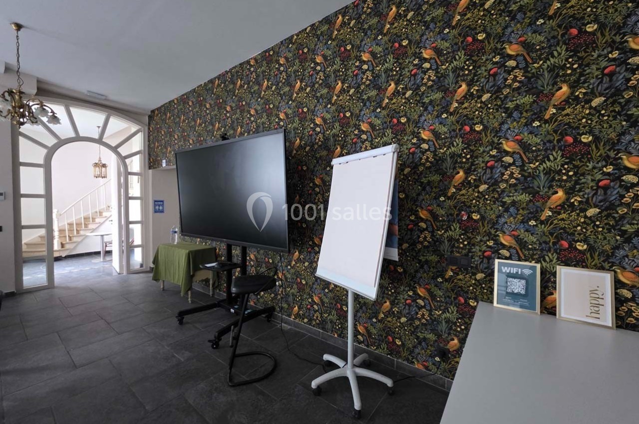 Salle équipée d'un écran, d'un chevalet avec tableau blanc et d'une table, décorée d'un papier peint à motifs floraux.