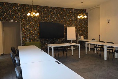 Salle de réunion lumineuse avec de longues tables blanches, chaises noires, écran mural et tableau blanc.