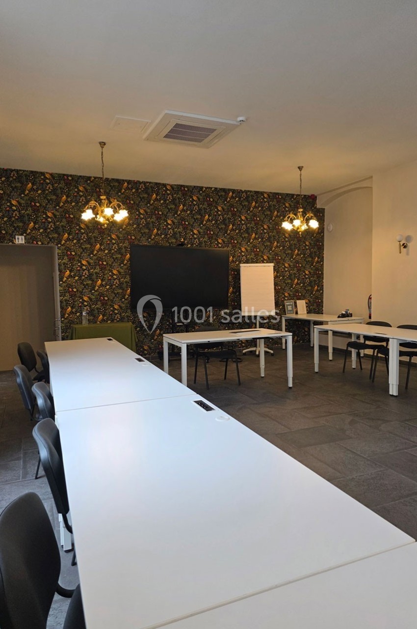 Salle de réunion lumineuse avec de longues tables blanches, chaises noires, écran mural et tableau blanc.