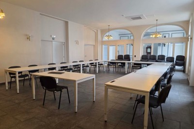 Salle de réunion lumineuse avec de longues tables blanches, chaises noires, écran mural et tableau blanc.