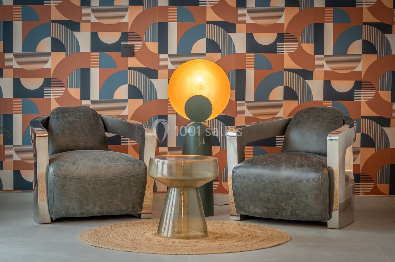 Deux fauteuils gris entourent une table basse en verre, éclairés par une lampe sur pied devant un mur à motifs géométriques.