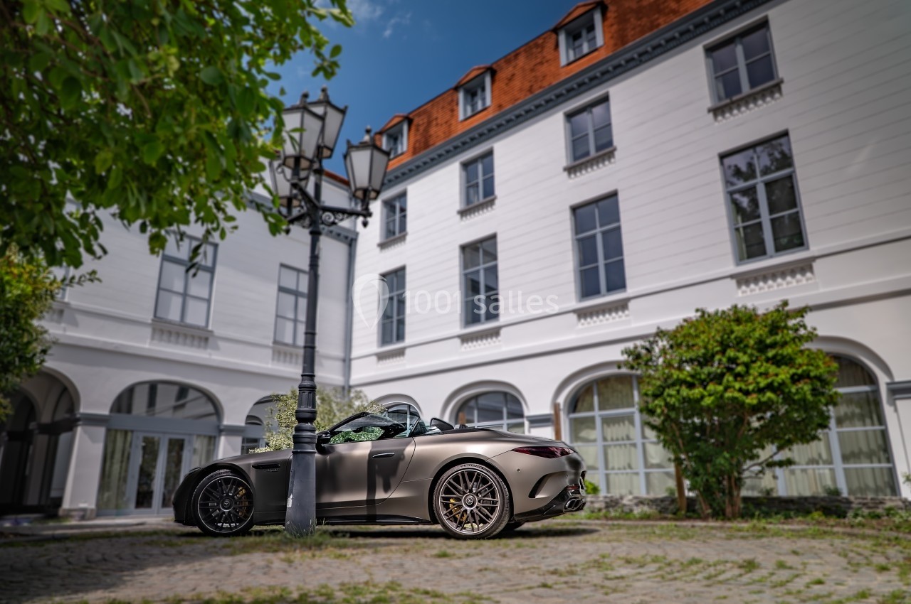 Voiture décapotable grise stationnée devant un bâtiment blanc à plusieurs étages, entourée de verdure.