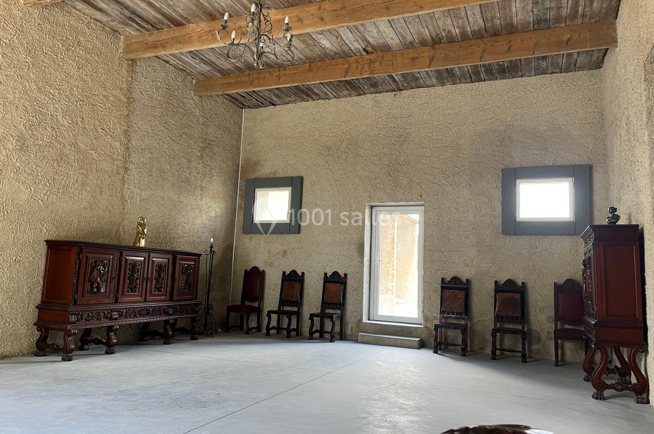 Salle spacieuse avec murs en pierre, plafond en poutres de bois, meubles anciens et chaises alignées contre le mur.