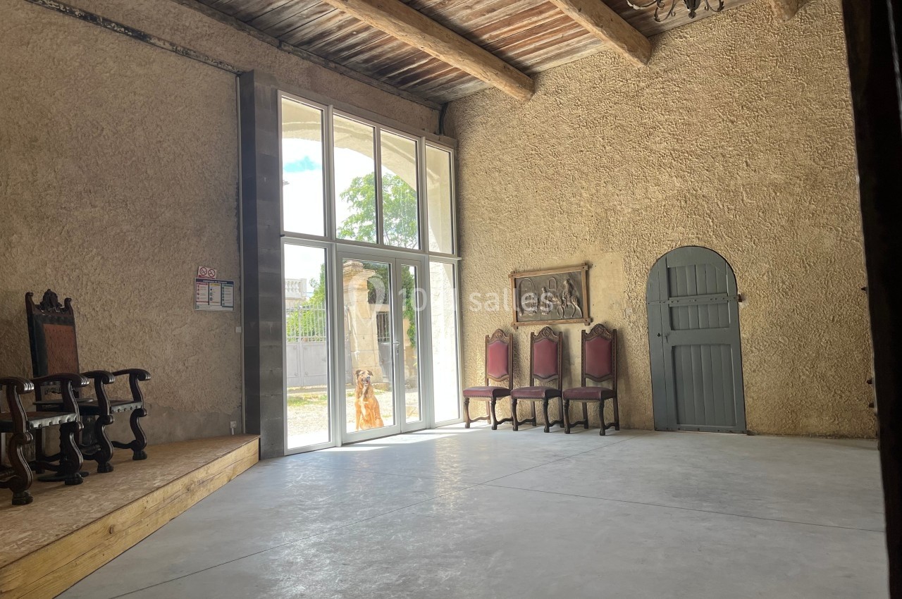 Salle avec murs en pierre, plafond en poutres apparentes, grandes fenêtres, chaises anciennes et porte en bois arrondi.