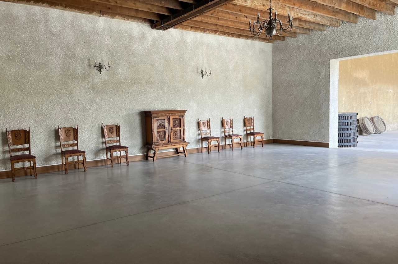 Salle vide avec sol en béton, plafond en poutres apparentes, chaises alignées contre un mur texturé et un buffet en bois.