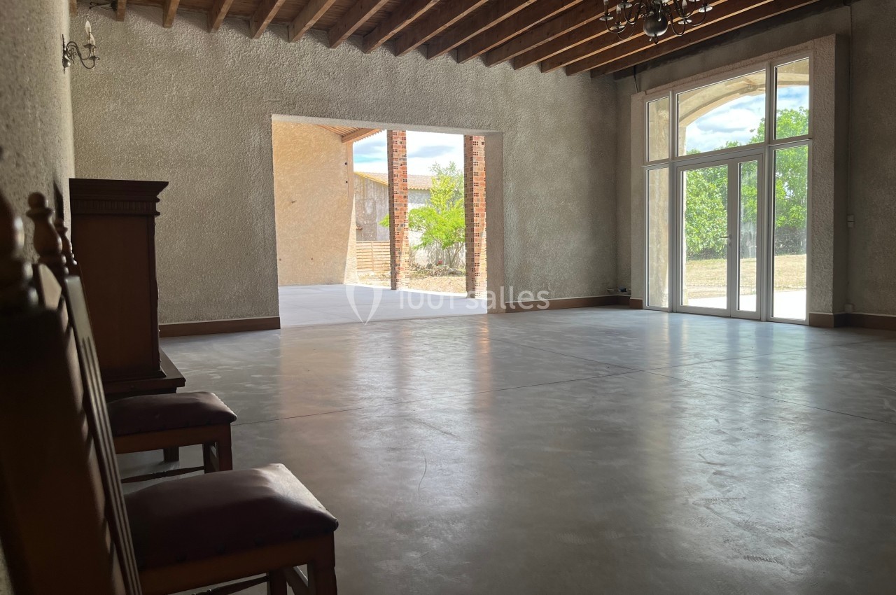 Salle vide avec sol en béton, plafond en bois, grandes fenêtres donnant sur un extérieur ensoleillé avec végétation.