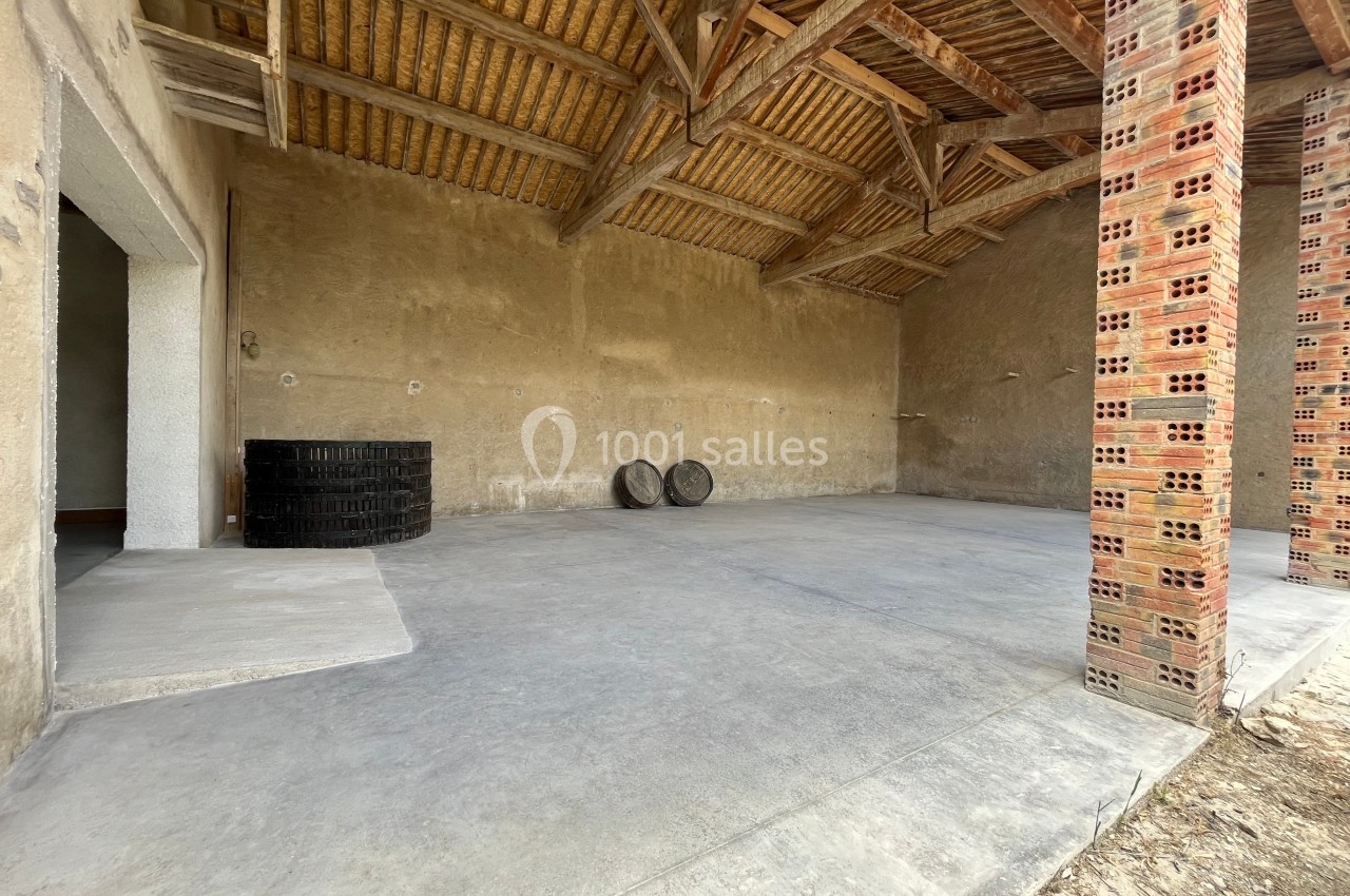 Intérieur d'un bâtiment avec sol en béton, murs en pierre, toit en bois et deux tonneaux posés au fond.