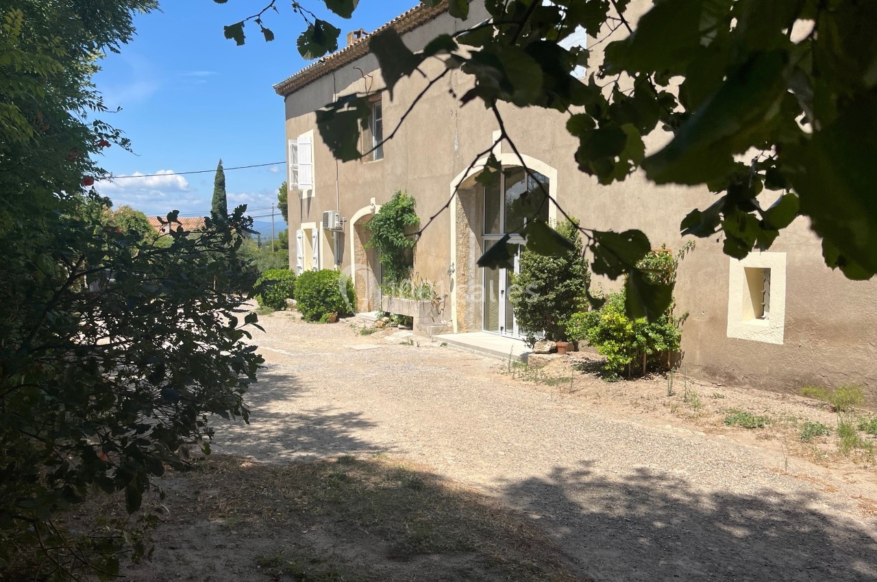 Façade d'une maison en pierre beige entourée de végétation, avec une allée en gravier sous un ciel bleu.
