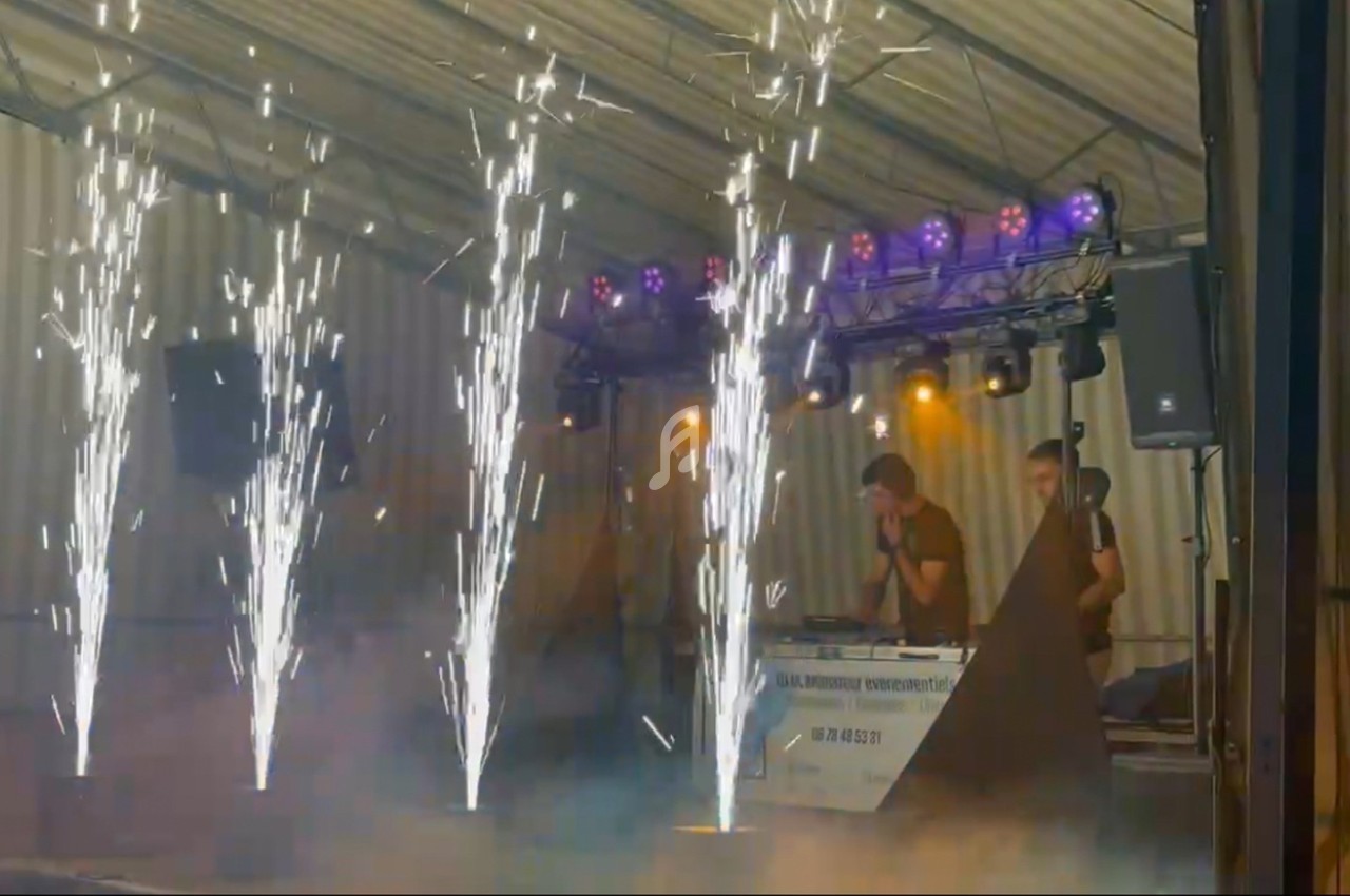 Effets pyrotechniques devant une scène avec des lumières colorées et un DJ en arrière-plan.