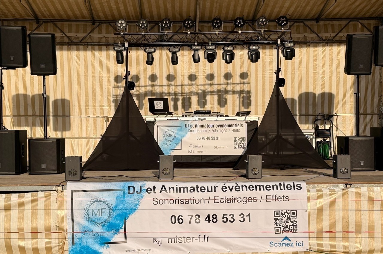 Scène extérieure équipée de matériel de sonorisation, éclairage et effets, avec une bannière promotionnelle au premier plan.