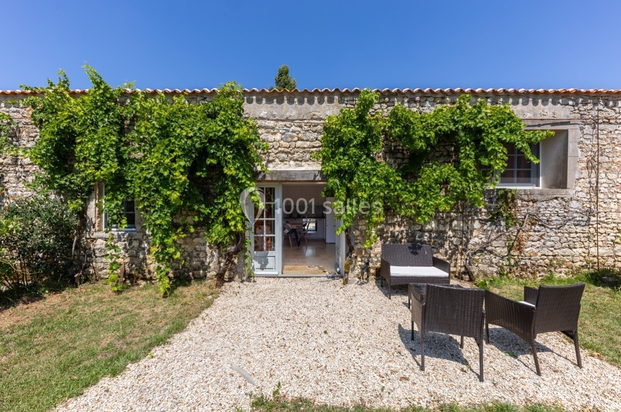 Façade en pierre d'une maison avec vigne grimpante, terrasse en gravier et mobilier de jardin en rotin.