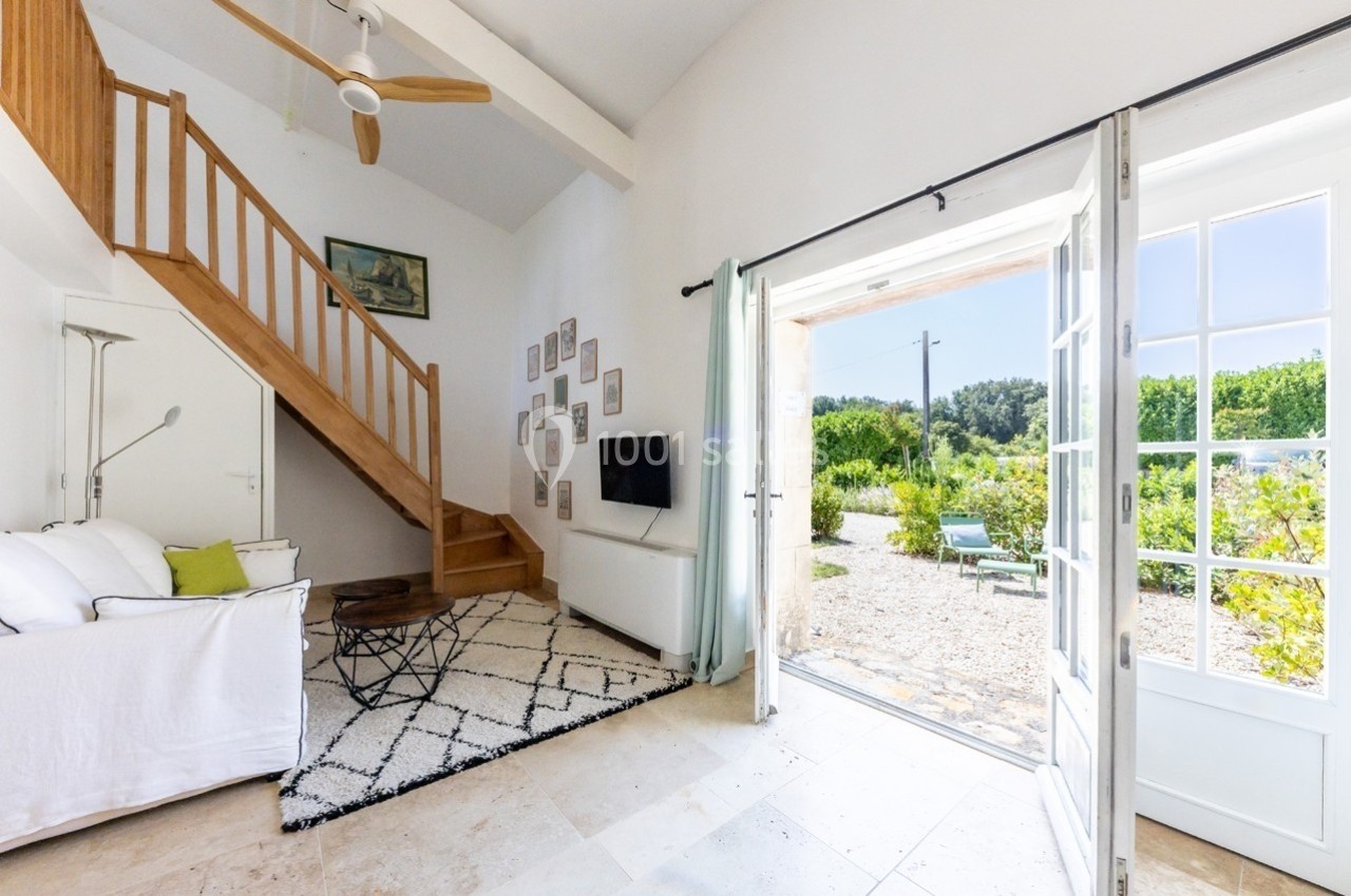 Salon lumineux avec canapé, escalier en bois, télévision, tapis géométrique et porte ouverte sur un jardin verdoyant.