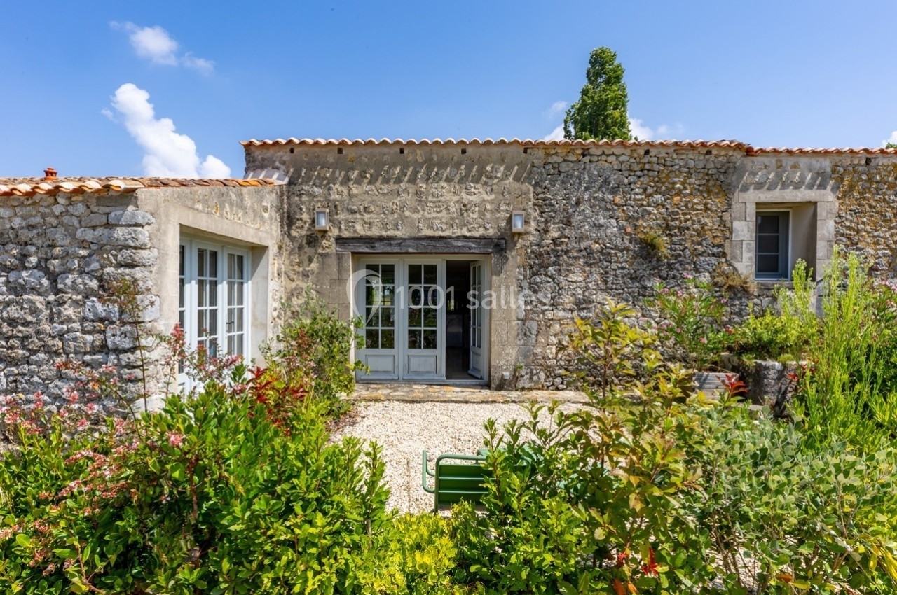 Façade en pierre d'une maison avec toit en tuiles, entourée de végétation et d'un jardin fleuri sous un ciel bleu.