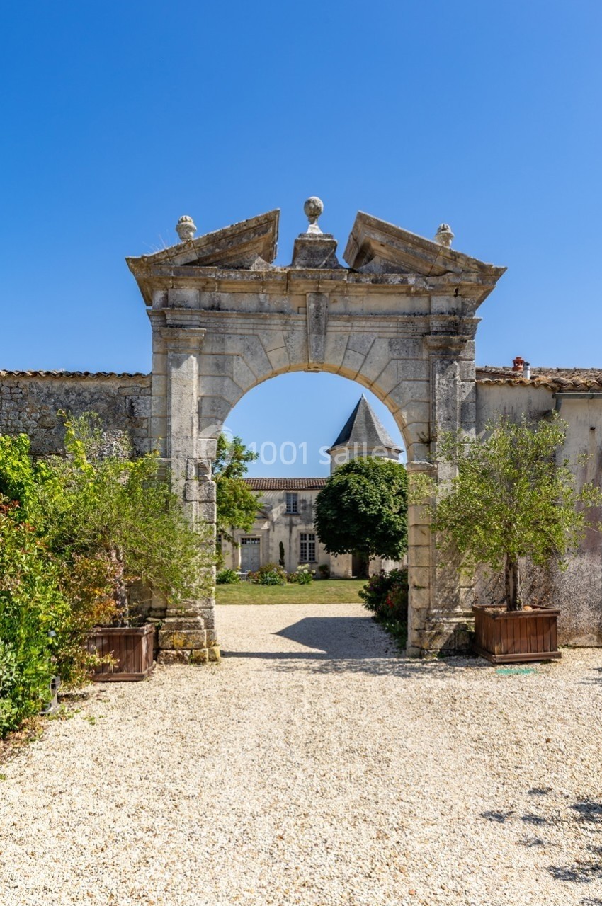 Arc en pierre ancienne encadrant une cour gravillonnée avec des bâtiments historiques et un clocher en arrière-plan.