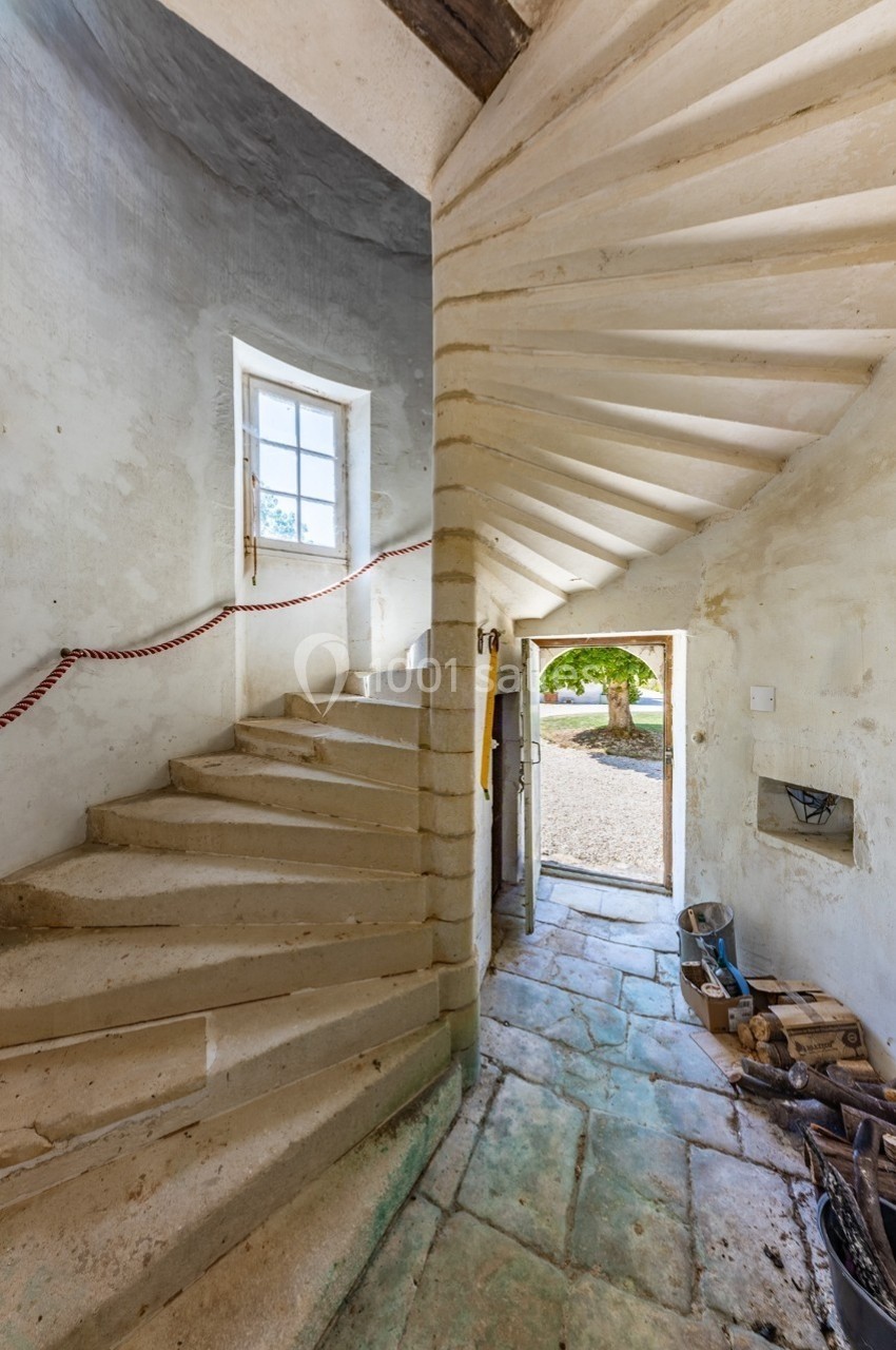 Escalier en pierre courbé dans un intérieur ancien, avec une porte ouverte donnant sur une cour ensoleillée.