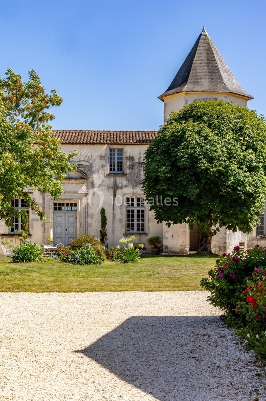 Façade d'une maison ancienne avec tour, entourée d'arbres, d'un jardin fleuri et d'une allée en gravier.