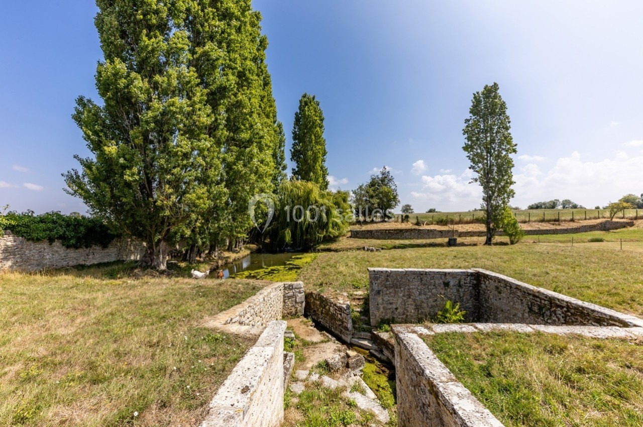 Vue d'un paysage rural avec des arbres, un petit cours d'eau et des structures en pierre dans un champ ensoleillé.