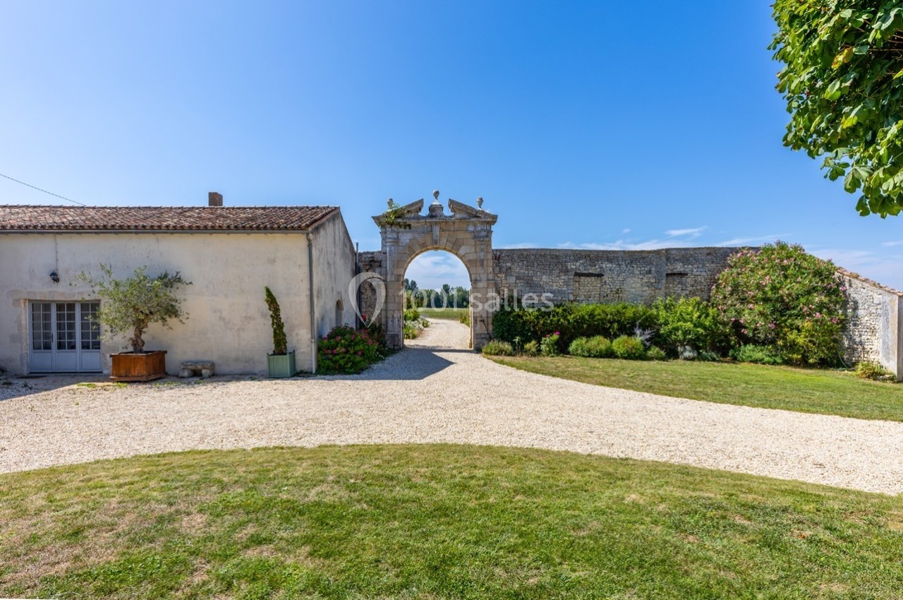 Entrée d'une propriété avec une arche en pierre, entourée de bâtiments anciens et d'un jardin verdoyant.