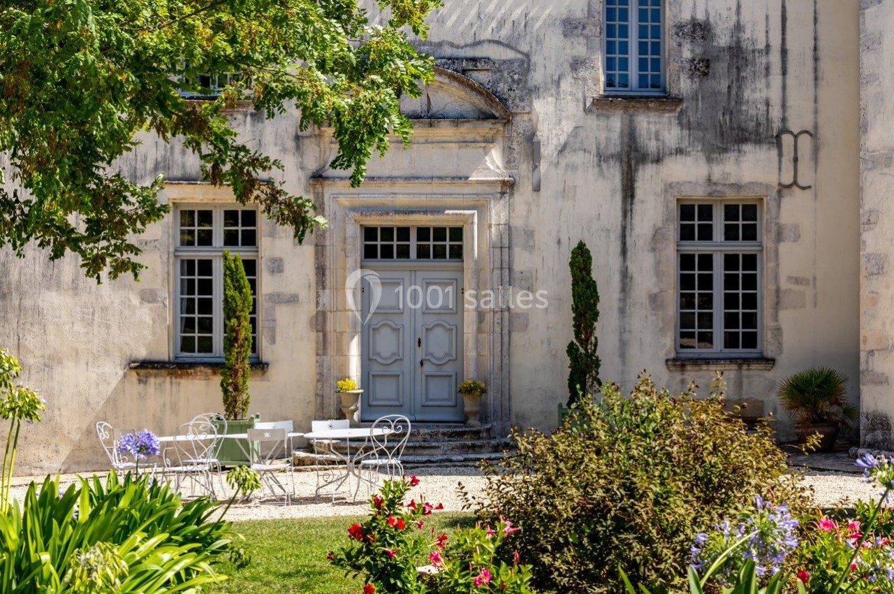 Façade d'un bâtiment ancien avec porte centrale, fenêtres symétriques et mobilier de jardin entouré de végétation fleurie.