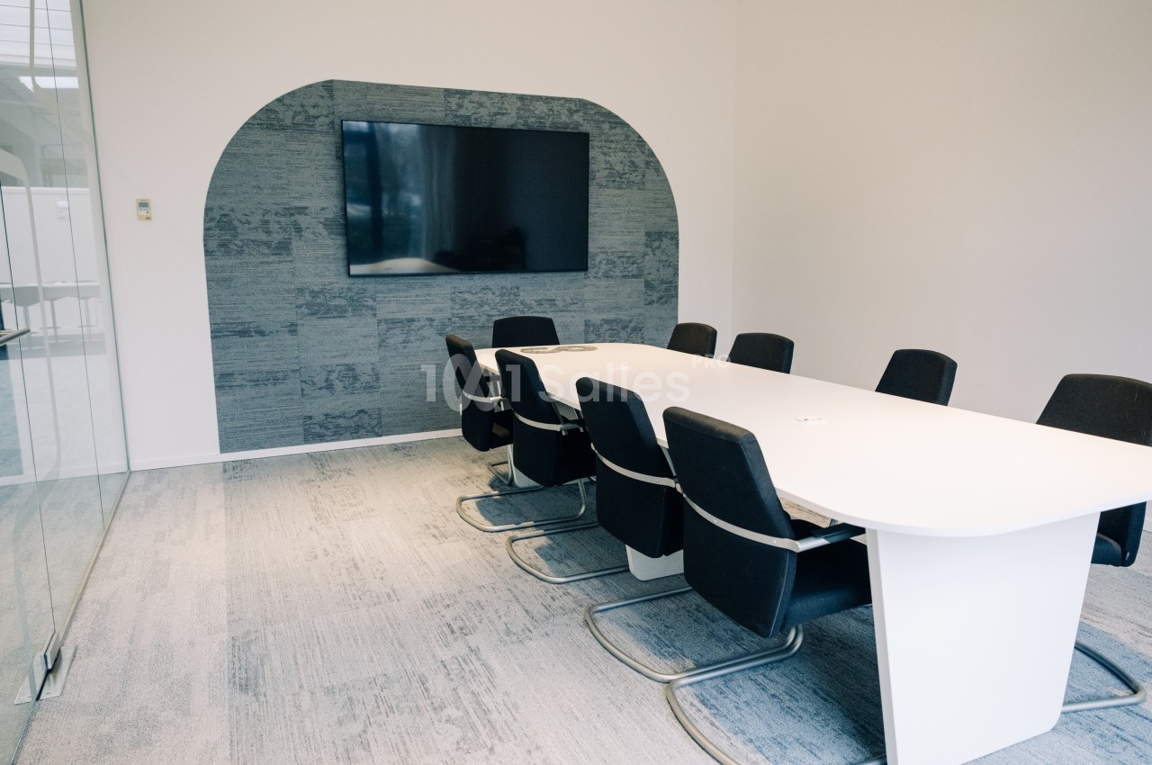 Salle de réunion moderne avec une grande table blanche, des chaises noires et un écran mural.