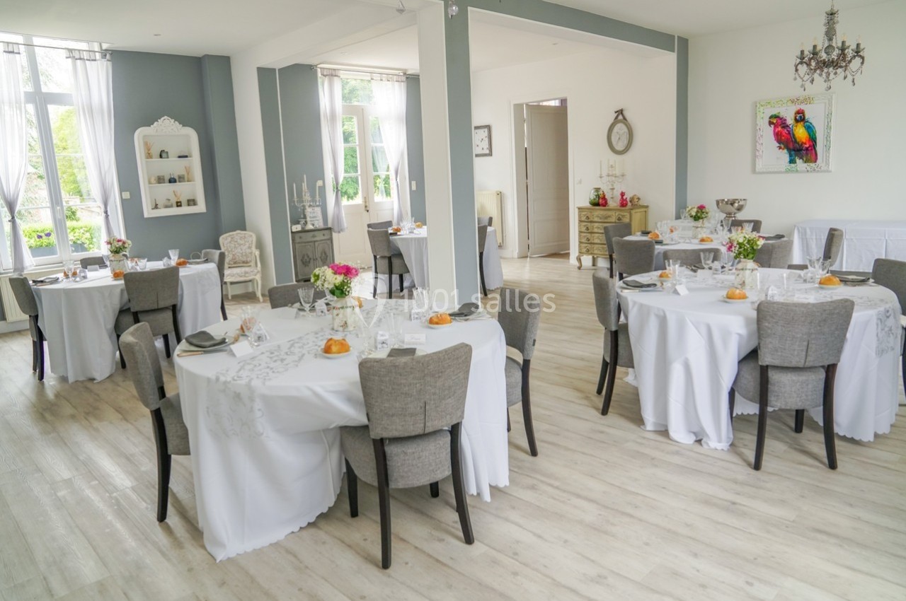 Salle lumineuse décorée pour un repas, avec des tables rondes dressées et des chaises grises.