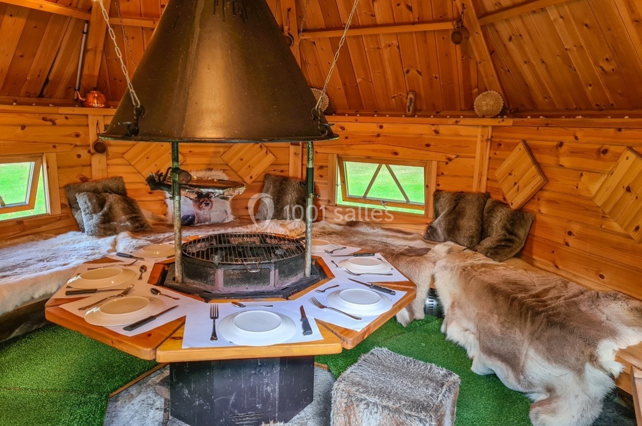 Intérieur d'un chalet en bois avec un barbecue central, des assises recouvertes de fourrures et une table dressée.