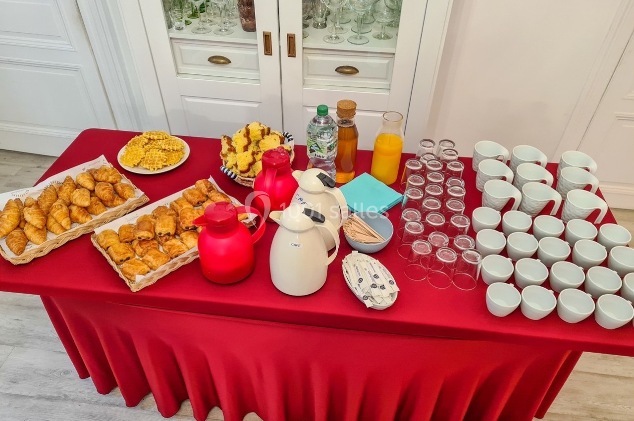 Table avec nappe rouge présentant viennoiseries, fruits, boissons chaudes et froides, tasses et verres alignés.