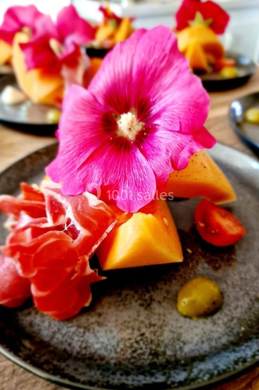 Assiette garnie de jambon cru, morceaux de melon, tomates cerises et une fleur comestible rose en décoration.