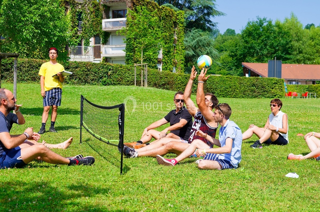 Des personnes assises sur l'herbe jouent au volley avec un filet, dans un parc ensoleillé entouré de verdure.