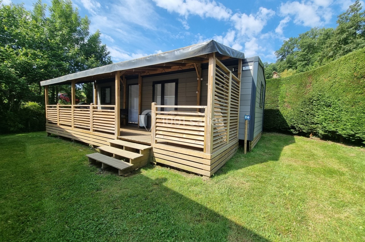 Chalet en bois avec terrasse couverte, entouré de pelouse et de haies, sous un ciel bleu partiellement nuageux.