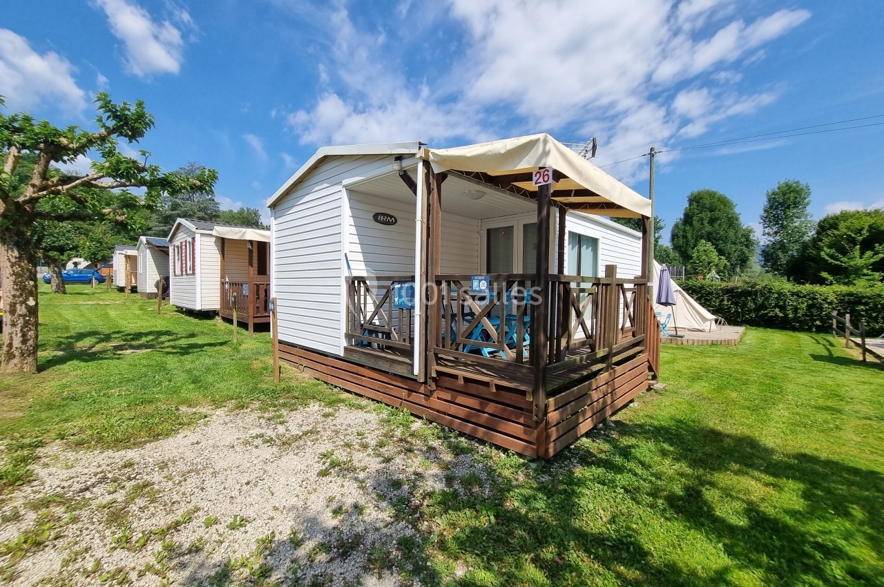 Mobil-home blanc avec terrasse en bois, situé dans un espace verdoyant sous un ciel ensoleillé.