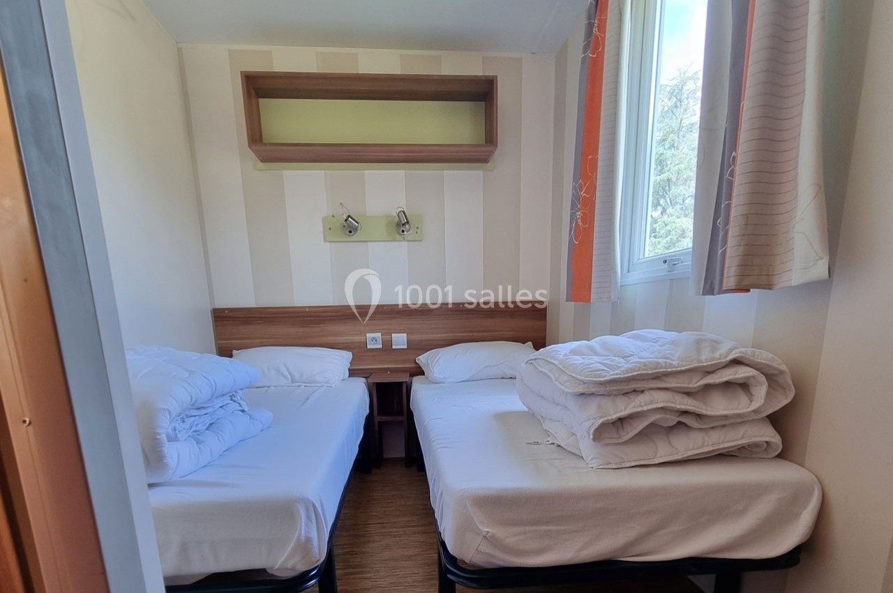 Chambre lumineuse avec deux lits simples, draps pliés, étagère murale et fenêtre donnant sur l'extérieur.
