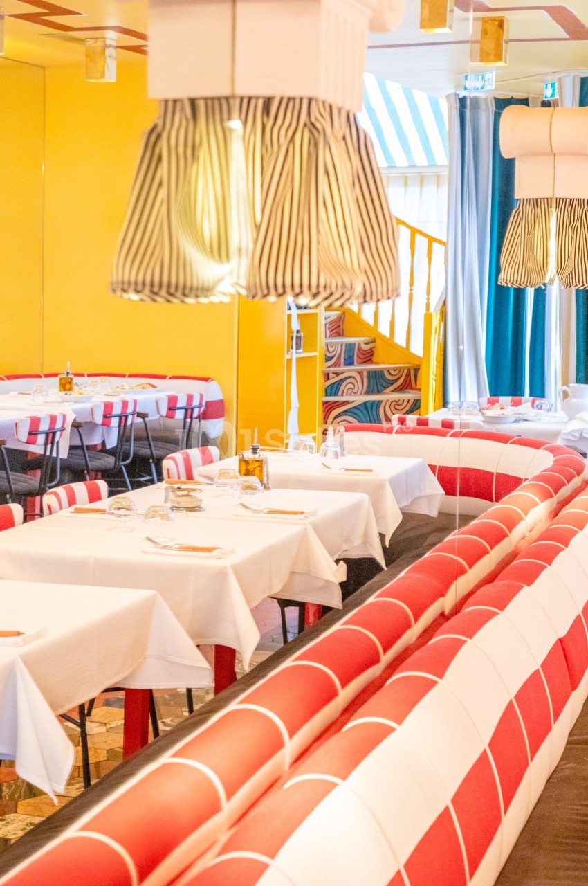 Salle de restaurant colorée avec banquettes rouges et blanches, nappes blanches et luminaires suspendus.
