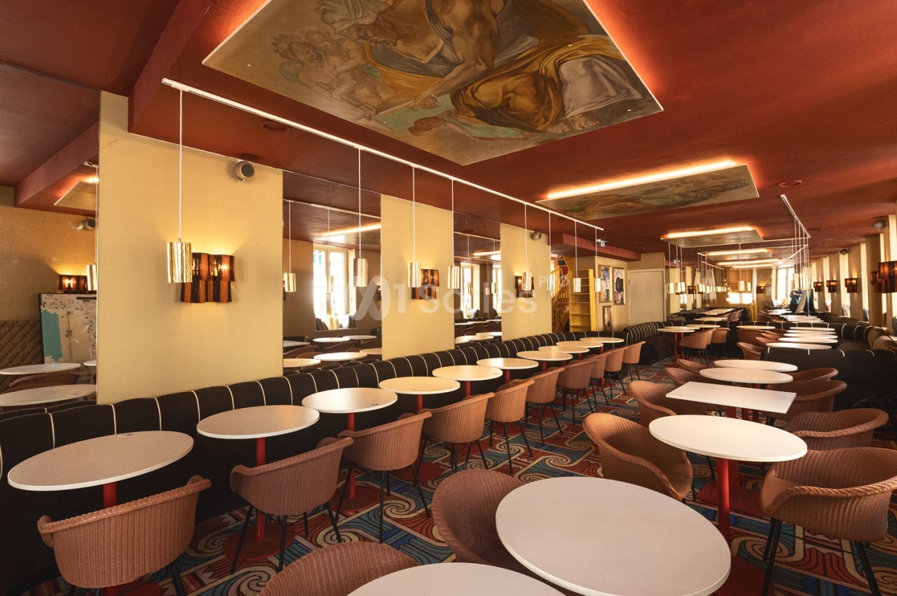 Salle intérieure avec tables rondes blanches, fauteuils marron et banquettes noires, décor rouille aux néons chauds.