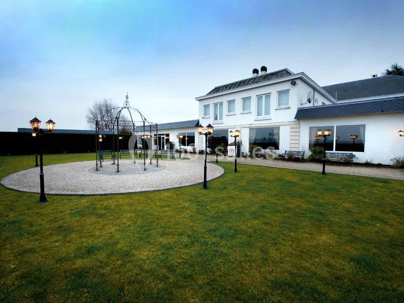 Grande maison blanche avec jardin, pelouse bien entretenue, lampadaires et une structure métallique circulaire au centre.